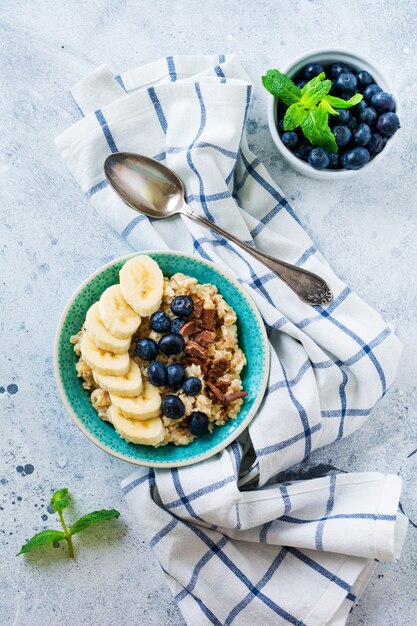 Haferbrei mit Banane, Schokolade und frischer Blaubeere in einer Schüssel auf einer hellgrauen Schiefer-, Stein- oder Betonoberfläche