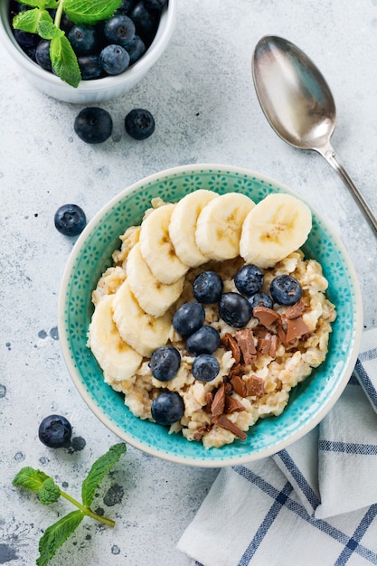 Haferbrei mit Banane, Schokolade und frischer Blaubeere in einer Schüssel auf einer hellgrauen Schiefer-, Stein- oder Betonoberfläche