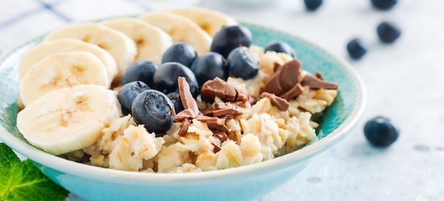 Haferbrei mit Banane, Schokolade und frischer Blaubeere in einer Schüssel auf hellgrauem Schiefer-, Stein- oder Betonhintergrund. Ansicht von oben.