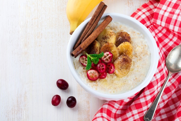 Haferbrei mit gebackenen Bananen, Zimt, Orange und Preiselbeeren in einer Keramikschale auf einer hellen Oberfläche. Draufsicht. Selektiver Fokus.