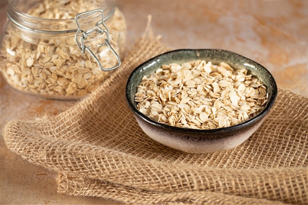 Haferflocken-Müsli in einer handgefertigten Keramikschale auf Sackleinen auf einem beigen Steintisch, ein Glasgefäß mit Haferflocken