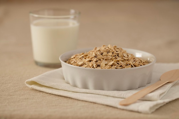 Haferflocken-Müsli in einer weißen Keramikschale auf einer Leinentischdecke, auf dem Tisch ein Glas Milch und ein Löffel