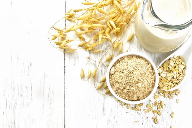 Hafermehl in einer Schüssel, Milch in einem Krug, Haferflocken im Löffel, Haferstiele auf Holzbretthintergrund von oben