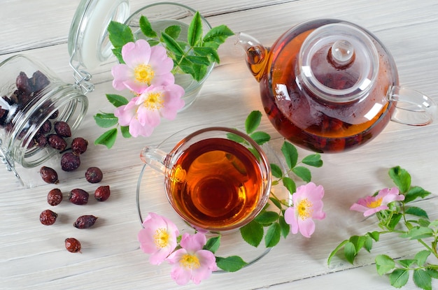 Hagebuttenblumen und eine Tasse Tee mit Hagebutten auf weißem Holztisch. Ansicht von oben.