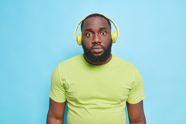 Halblange Aufnahme eines überraschten Mannes mit dickem Bart reagiert auf etwas Erstaunliches, das in einem lässigen T-Shirt gekleidet ist, hört Musik über Kopfhörer und hört schockierende Nachrichten einzeln über blauer Wand?