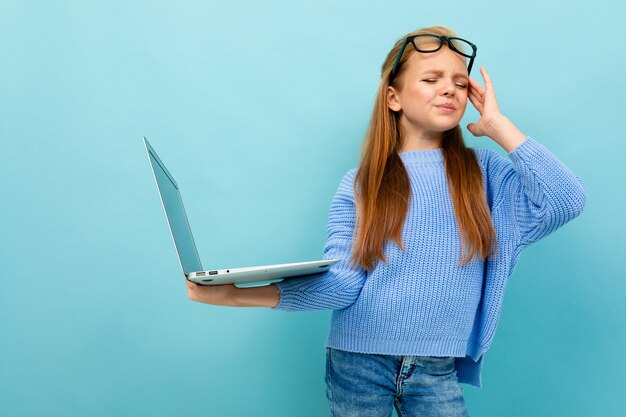 Halblanger Schuss eines Schulmädchens auf einem blauen Hintergrund mit müden Augen, Gläser auf ihrer Stirn in den Händen eines Laptops