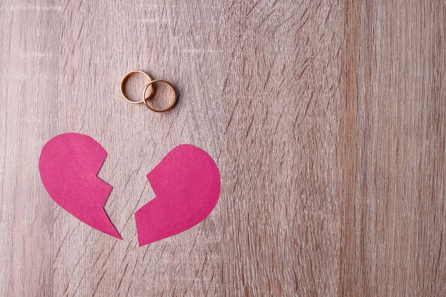 Halbteile von zerrissenen roten Papierherzen und Hochzeitsringen auf einem Holztisch