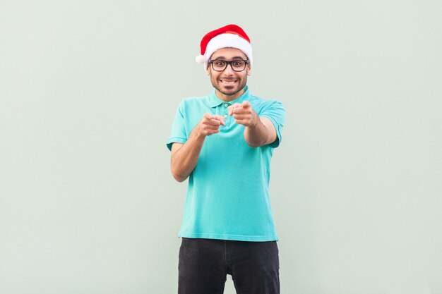 Hallo du! Sein Weihnachtsmann! Bärtiger Mann in Weihnachtsmütze und Brille, der mit dem Finger zeigt und mit einem zahnigen Lächeln in die Kamera schaut. Auf grauem Hintergrund. Innenaufnahme, Studioaufnahme