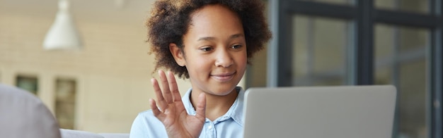 Hallo fröhliches Teenager-Schulmädchen, das seinen Laptop benutzt und Hallo winkt, während es mit Freunden oder Lehrern chattet