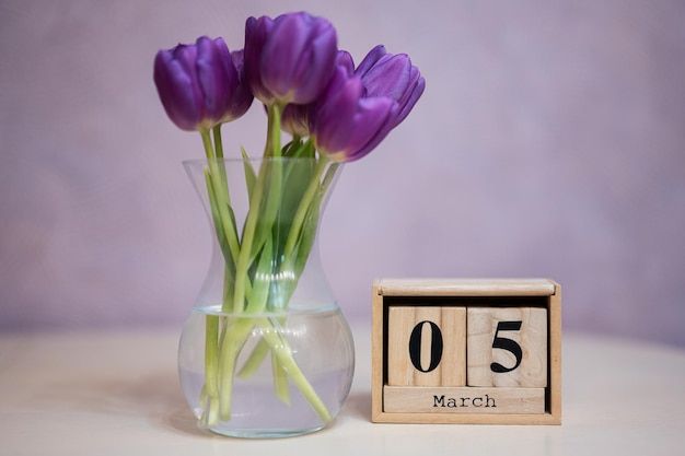 Hallo Frühling Holzkubischer Kalender mit Datum vom 5. März, umgeben von einem Strauß violetter Tulpen mit grünen Blättern in Glasvase