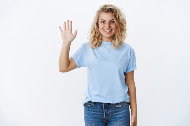 Hallo Ich heisse. Porträt einer freundlichen und charismatisch lächelnden jungen, lockigen Blondine mit blauen Augen, die die Handfläche in Hallo heben und die Geste begrüßen, um neue Leute kennenzulernen, die einladend grinsen