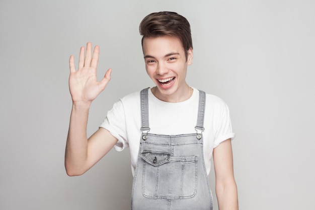 Hallo, schön, dich zu sehen. Fröhlicher erstaunter junger brünetter Mann im lässigen Stil, Denim-Overalls, die stehen und die Kamera betrachten und mit einem zahnigen Lächeln begrüßen. Indoor-Studioaufnahme, auf grauem Hintergrund isoliert.