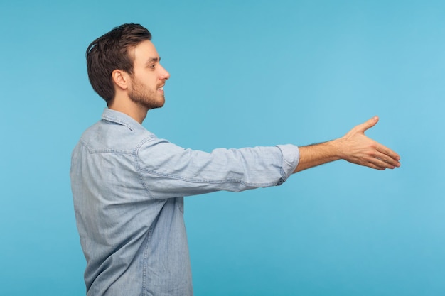 Hallo, schön, Sie kennenzulernen Seitenansicht eines freundlich lächelnden, gutaussehenden Mannes, der im Arbeiter-Jeanshemd die Hand ausstreckt und Handschlag beim Vorstellungsgespräch im Innenstudio anbietet, isoliert auf blauem Hintergrund