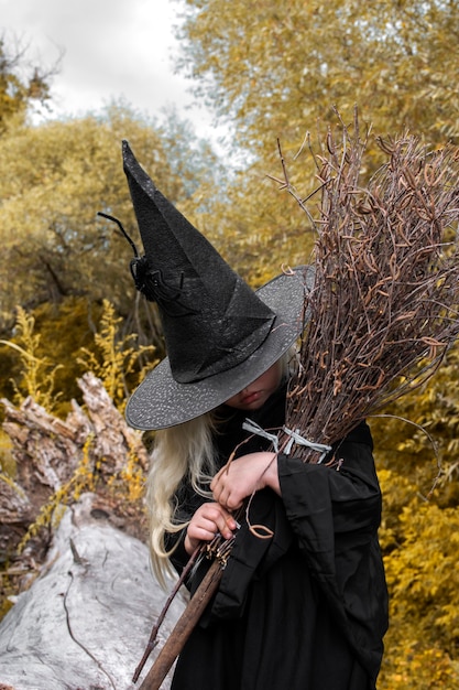 Halloween y brujas. Chica de sombrero negro con una escoba en sus manos en el bosque de otoño