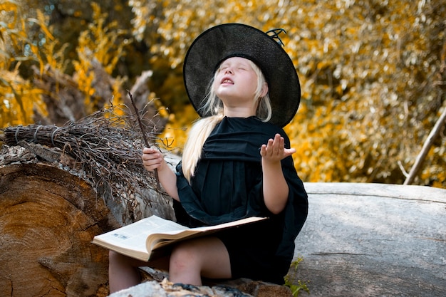 Halloween y brujas. Niño disfrazado de bruja. El niño está sentado en un árbol con un libro y una varita mágica. Otoño, bosque