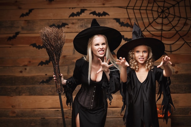 Halloween concept mãe alegre e sua filha em fantasias de bruxa comemorando o halloween posando com abóboras curvas sobre morcegos e teia de aranha no fundo do estúdio de madeira