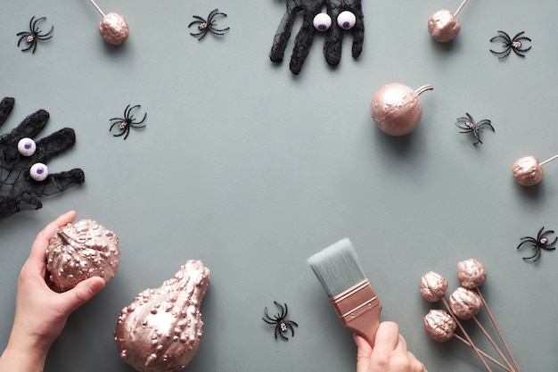 Halloween criativo plana colocar no papel cinza com cópia-espaço. Moldura feita de abóboras douradas rosa, luvas de malha preta com olhos de chocolate, mão com pincel e algumas aranhas negras.