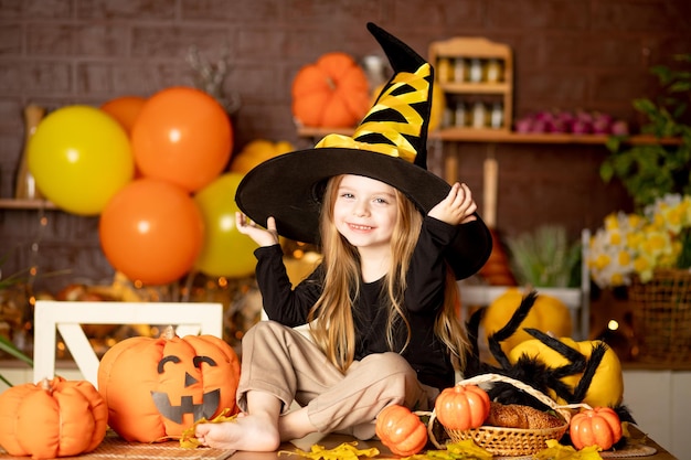 Halloween freut sich ein süßes lächelndes Kindermädchen in einem Hexenkostüm in einer dunklen Küche mit Kürbissen und einer großen Spinne und festlichem Dekor während der Halloween-Feier