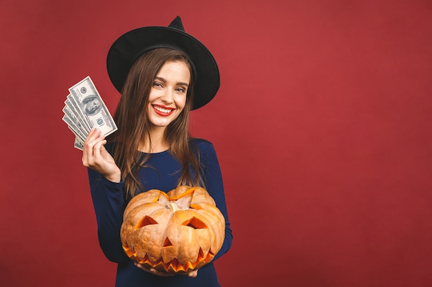 Halloween-Hexe mit einem geschnitzten Kürbis und Geld - lokalisiert auf rotem Hintergrund. Emotionale junge Frau im Halloween-Kostüm.