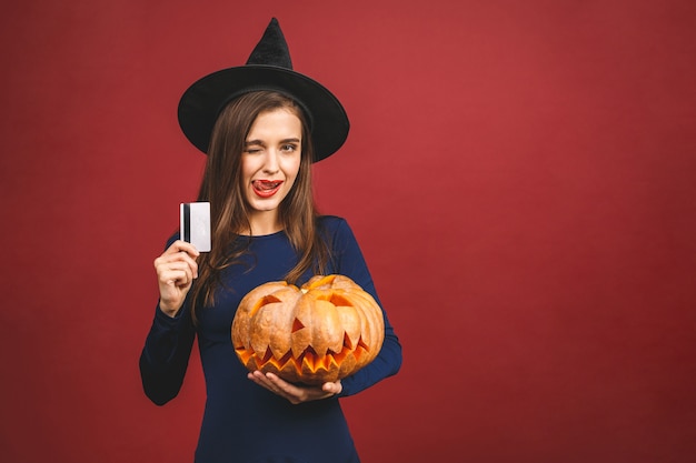 Halloween-Hexe mit einem geschnitzten Kürbis und Kreditkarte - lokalisiert auf rotem Hintergrund. Emotionale junge Frau im Halloween-Kostüm.