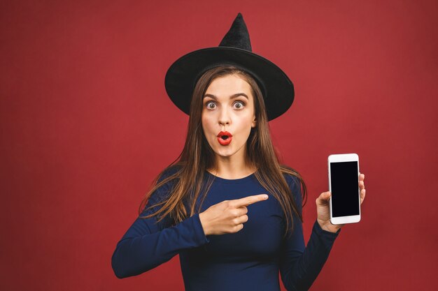 Halloween-Hexe mit Handy-Bildschirm - lokalisiert auf rotem Hintergrund. Emotionale junge Frau im Halloween-Kostüm. Halloween-Partygirl.