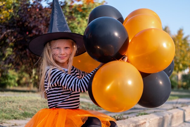 Halloween-Kinderporträt lächelndes Mädchen im Hexenhut mit den lustigen Kindern der orange und schwarzen Ballone in