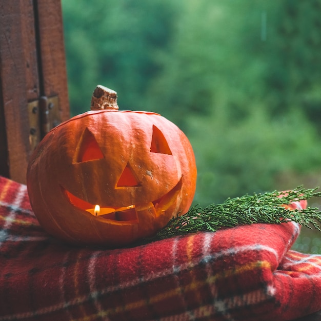 Halloween-Kürbis auf einem gemütlichen Fensterbrett mit einem roten Plaid. Ganzer Kürbis und Wunderkerze im Freien. Fröhliches Halloween! Der Herbst ist gemütlich. Regen draußen.