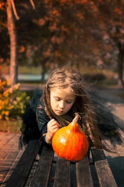 Halloween liegt ein süßes Mädchen mit langen Haaren auf einer Bank im Park und zeichnet ein lustiges Gesicht zum Kürbis