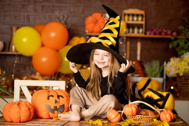 Halloween una linda niña sonriente con un disfraz de bruja en una cocina oscura con calabazas y una gran araña y una decoración festiva durante la celebración de Halloween se regocija