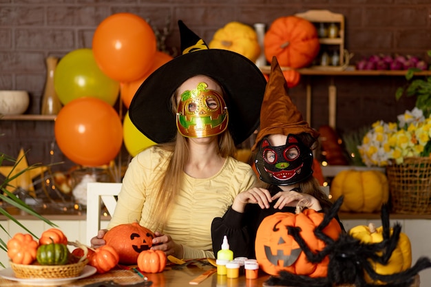 Foto halloween, mama und kindertochter in einem hexenkostüm und karnevalsmasken in einer dunklen küche mit kürbissen und einer großen spinne erschrecken mit den händen, lächeln, jubeln