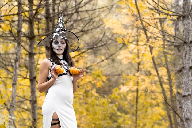 Halloween Menina linda em um vestido branco com maquiagem no rosto em um chapéu de bruxa posando na floresta de outono Ele segura duas pequenas abóboras nas mãos e sorri O dia dos mortos