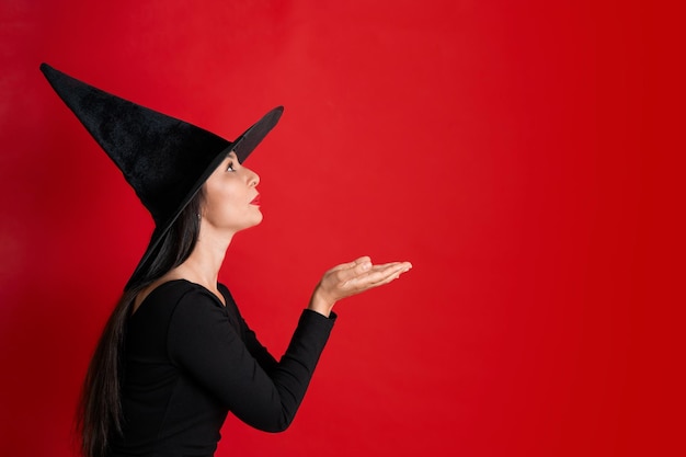 Halloween Una mujer hermosa con un disfraz de bruja negra y un sombrero está conjurando hace lugar mágico para el fondo rojo del texto