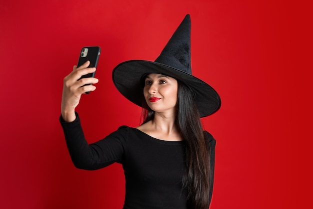 Halloween Una mujer hermosa joven en un traje de bruja y un sombrero toma un selfie en su teléfono Fondo rojo Lugar para el texto