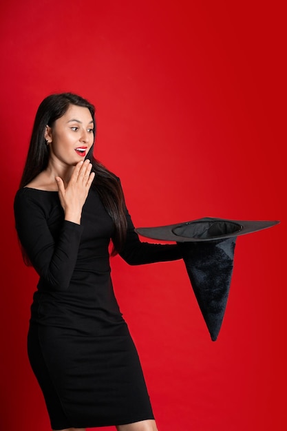 Halloween Una mujer hermosa en un negro sostiene un sombrero mágico en sus manos y reacciona emocionalmente Lugar para el texto de fondo rojo