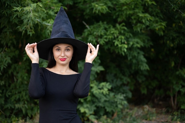 Halloween Una mujer joven y hermosa con un sombrero de bruja y un vestido negro en el parque Espacio para texto