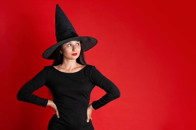 Halloween Una mujer joven y hermosa con un sombrero de bruja y un vestido negro sobre un fondo de color rojo Espacio para texto