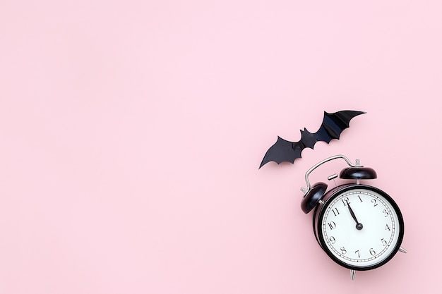 Halloween-Nacht-Konzept. Schwarzer Wecker und fliegende Fledermaus auf rosa Hintergrund. Kreative flache Lage, Draufsicht
