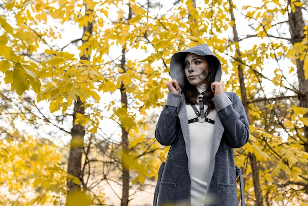 Halloween-Porträt Ein junges schönes Mädchen in einem weißen Kleid mit Make-up im Gesicht in einem Kapuzenmantel steht gruselig im Herbstwald Der Tag der Toten