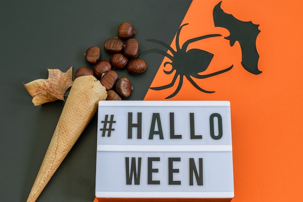 Halloween-Text auf schwarz-orangeem Papier mit Waffeleistüte mit Kastanien und dekorativer Fledermaus und Spinne. Flach liegen