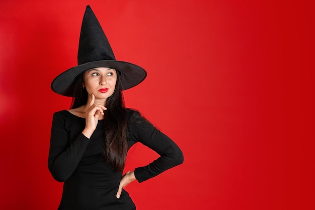 Halloween Uma jovem linda com um chapéu de bruxa e um vestido preto em um fundo vermelho Espaço para texto