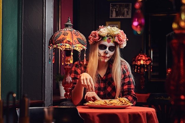 Halloween- und Muertos-Konzept. Junges blondes Mädchen mit untotem Make-up im Blumenkranz, das in einem mexikanischen Restaurant Nachos isst.