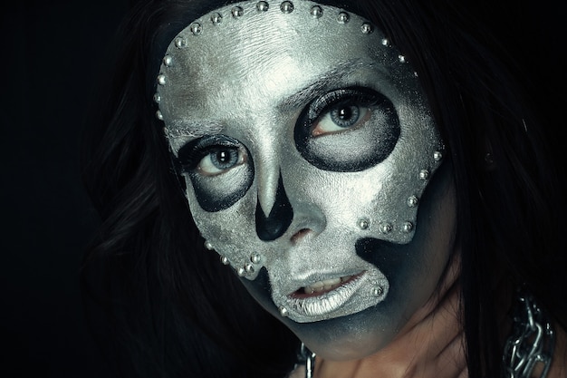 Foto halloween, vacaciones, estilo de vida, gente, belleza, concepto creativo - halloween y tema de maquillaje creativo: modelo de niña hermosa con cuerpo negro con pintura de calavera de máscara plateada sobre fondo oscuro en estudio