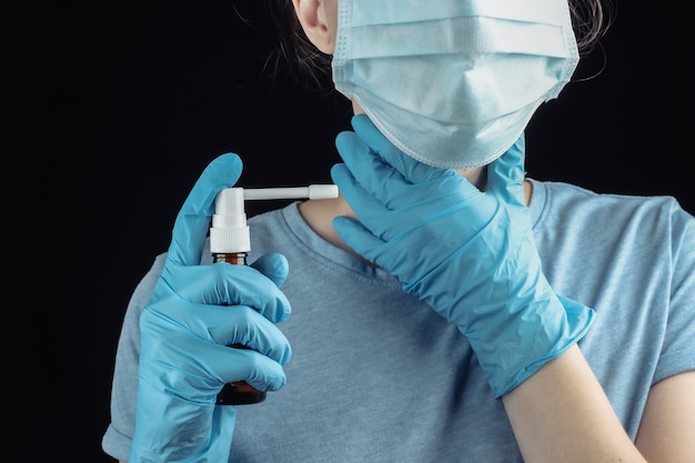 Halsentzündung. Symptome der Grippe. Frau in der medizinischen Maske und in den Handschuhen hält Flasche des Kehlensprays nah oben