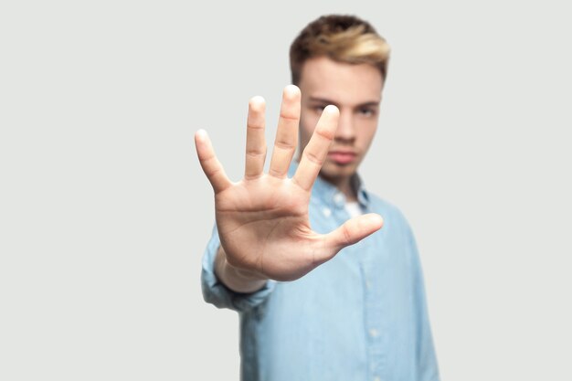 Halt. Porträt eines ernsten, gutaussehenden jungen Mannes in hellblauem Hemd, der die Kamera mit Wutgesicht betrachtet und die Stopphand zeigt. Innenstudio auf grauem Hintergrundkopierraum gedreht.