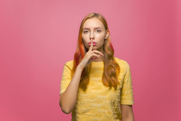 Halten Sie die Stille, junges, modernes Teenager-Mädchen, das eine stille Geste zeigt, die den Finger auf den Lippen auf dem rosa Studio hält