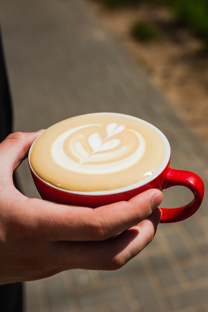 Halten Sie eine frisch zubereitete rote Tasse Cappuccino-Kaffee im Sonnenlicht