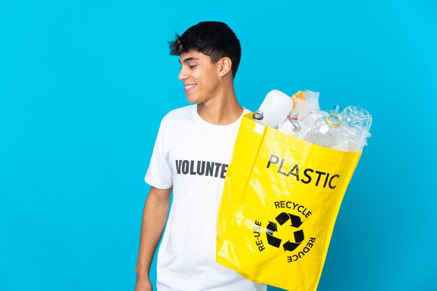 Halten Sie eine Tasche voller Plastikflaschen, um über blaue Wand zu recyceln, die zur Seite schaut und lächelt