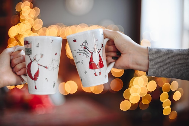 Halten Sie festliche Weihnachtstassen mit dem Weihnachtsmann auf dem Hintergrund des Weihnachtsraums mit verschwommenen Lichtern in Händen