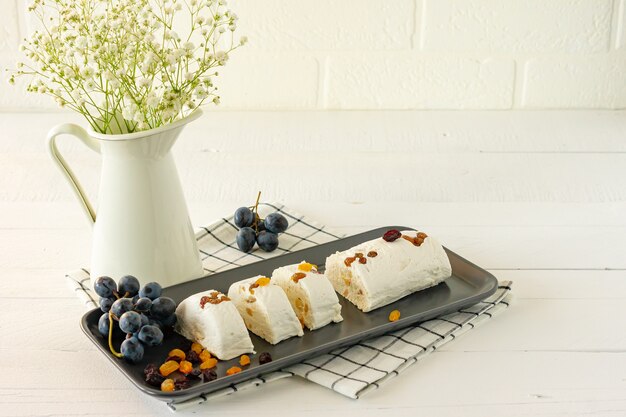 Halva fatiada com nozes e frutas secas em um prato servido com flores e frutas na mesa branca.