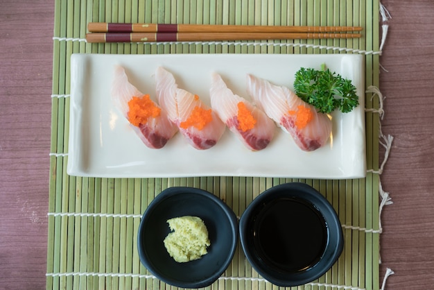 Hamachi sushi en plato blanco junto con salsa japonesa y decoración de hojas verdes.